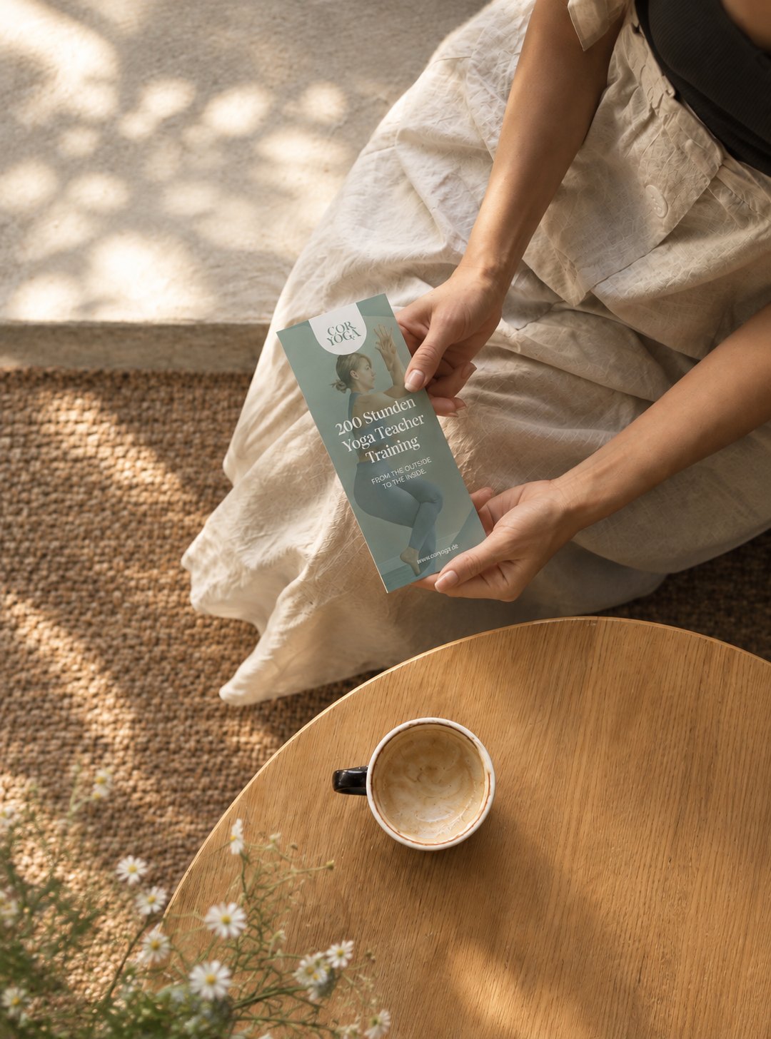 A woman holding a Coryoga flyer, seen from above, next to a coffee cup and daisies